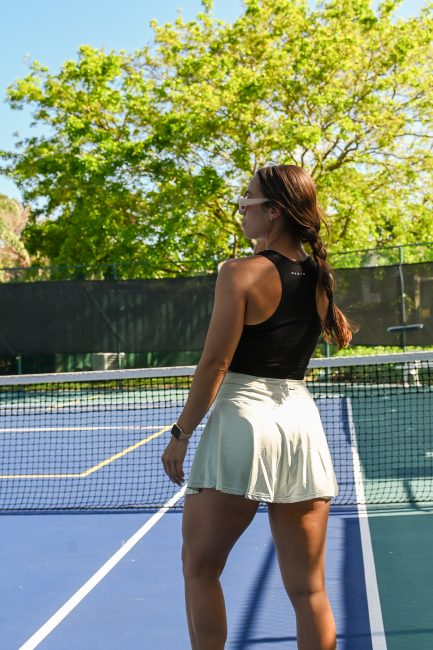 a girl is standing in front of a tennis net wearing a black tank top, green tennis skirt, sunglasses and white sneakers. 